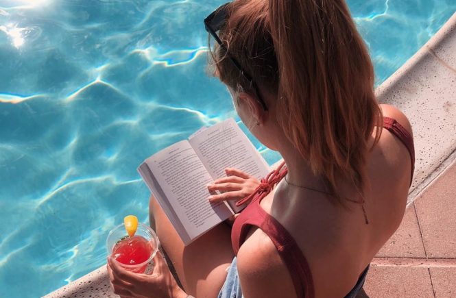 Blonde girl in a pink swimsuit sitting by the pool reading a book and drinking a vodka lemonade in Carlsbad, California.