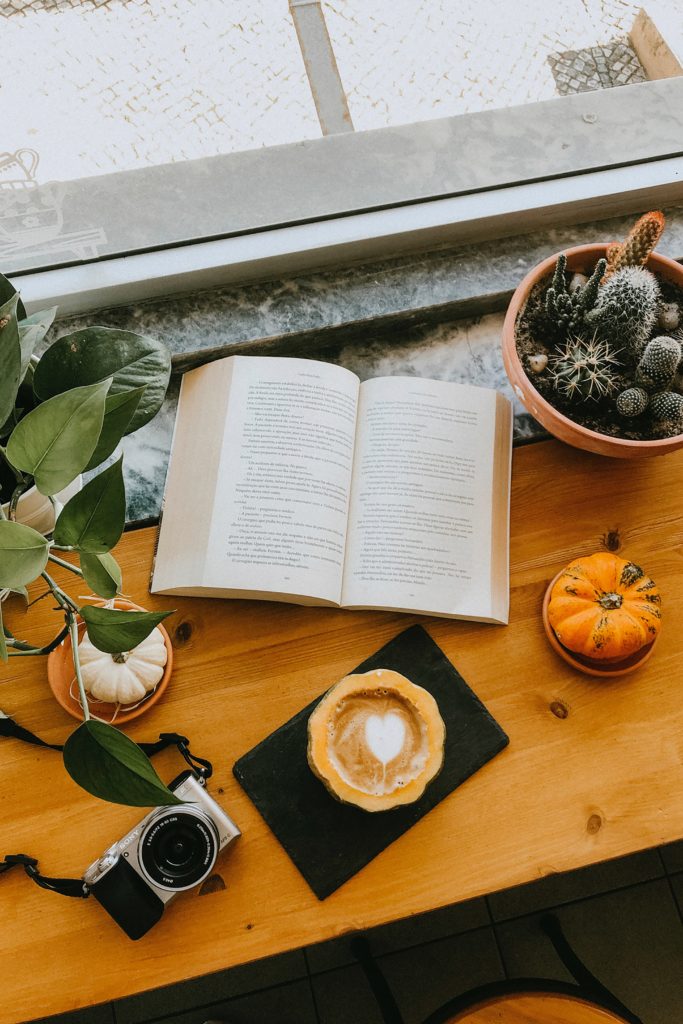 An amazing book next to a pumpkin and a pumpkin spice latte coffee on a table next to a camera.