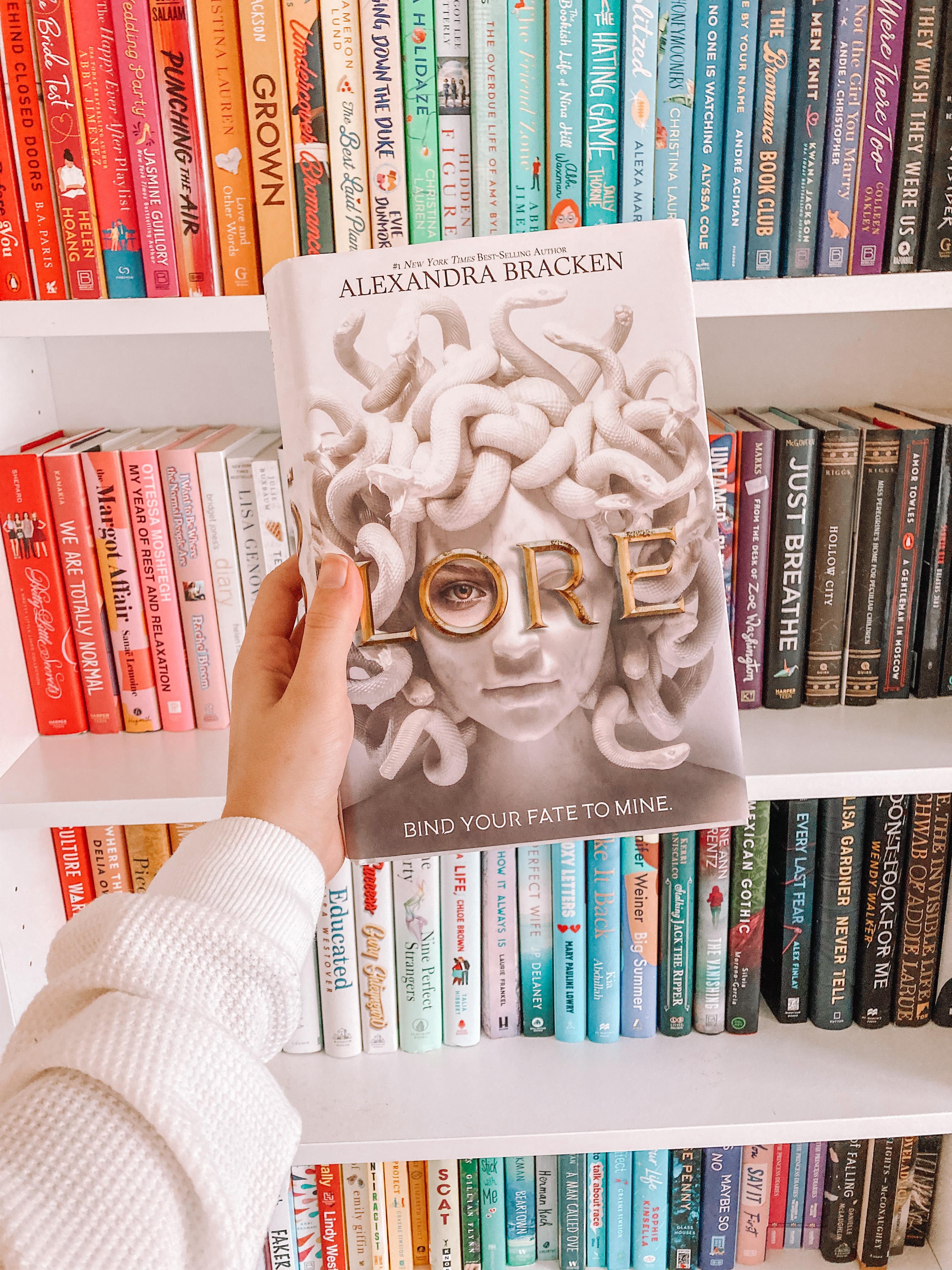 Book blogger holds a finished copy of Lore by Alexandra Bracken, a young adult fantasy novel, showing a Lore book review.